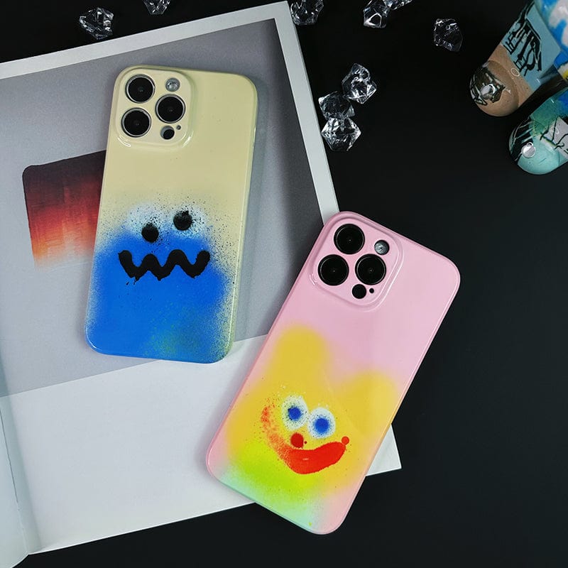 Two iPhones featuring cases from the Colorful Spray Art iPhone 16 Pro Max Case Set - Playful Faces in Vibrant Graffiti Style lie on an open magazine. The left case is cream and blue with a sad face, while the right has a pink and yellow smiling face. Small decorative crystals and a painted container are also visible in the background.