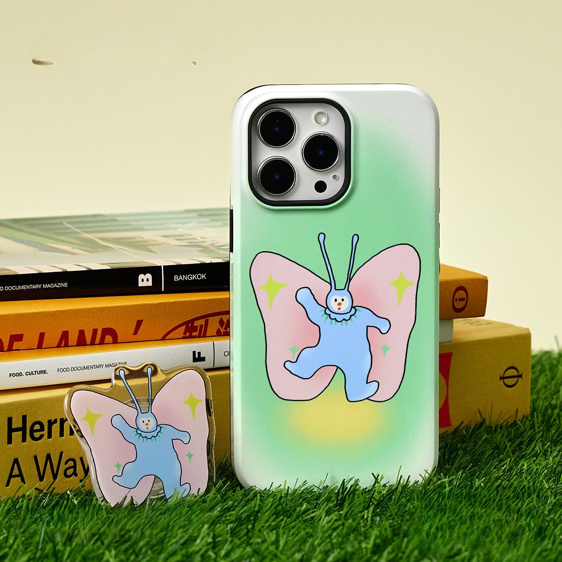 A Golden Sunset MagSafe iPhone 16 Pro Max case featuring an elegant gradient design and a sturdy ring stand stands upright on a patch of grass beside a matching accessory, with stacked books in the background.