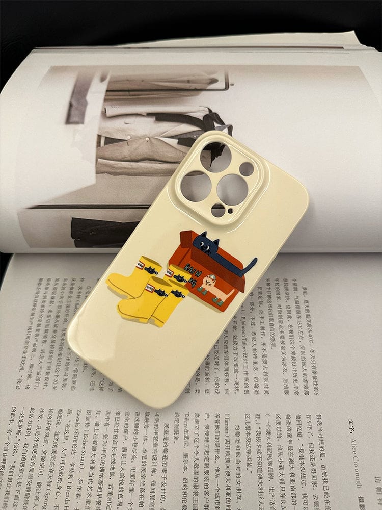 A beige Cute Cat in Rain Boots iPhone 16 Pro Max Case - Playful and Protective Design, featuring a colorful illustration of a black cat, yellow rain boots, and a small house, rests on top of an open magazine with Chinese text and monochrome images of fashion items.