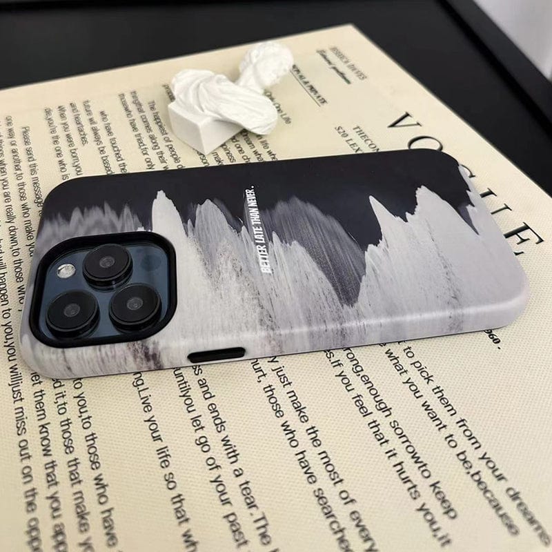 A phone with a rugged, mountain-inspired case sits on an open magazine. The Monochrome Waves iPhone 16 Pro Max case features a black and white mountainous design with the words "Better Late Than Never" printed on it. White earphones are placed near the top left of the phone.