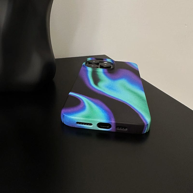 A smartphone with an Iridescent Waves iPhone 16 Pro Max Case - Sleek and Futuristic Design with Full Protection, featuring a colorful abstract pattern in shades of blue, green, and purple, lies on a black surface. Part of a black object is visible on the left side of the image. The phone's camera lenses are visible at the top.