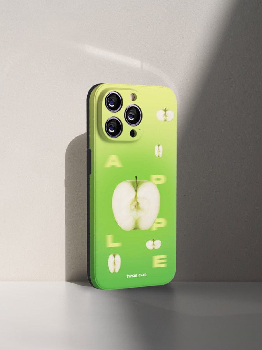 A smartphone stands upright on a white surface against a light gray background. The Green Apple Fresh iPhone 16 Pro Max Case, featuring a vibrant gradient design with images of sliced apples and letters that spell "APPLE" scattered among the apple slice images, protects the phone. The phone has three camera lenses arranged in a triangle.