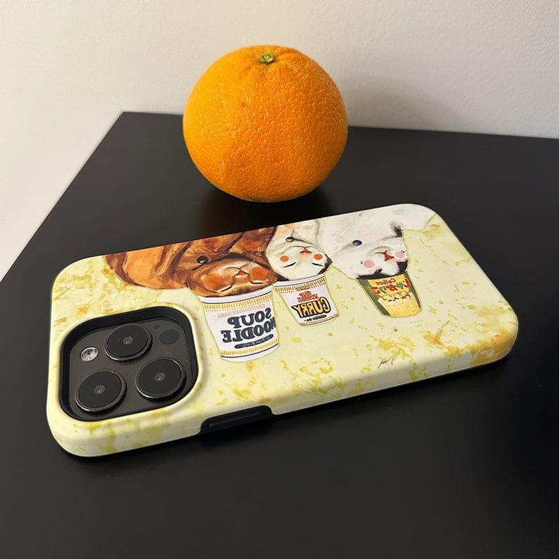 A smartphone with an Adorable Cats & Cup Noodles iPhone 16 Pro Max case, featuring a cute and quirky design, rests on a dark surface. In the background, an orange is placed against a light-colored wall.