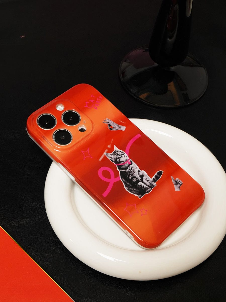 An iPhone 16 Pro Max encased in the Cat Vibes iPhone 16 Pro Max Case, featuring a playful orange gradient design adorned with various cat stickers and drawn elements like a pink bow, hand gestures, and stars. The smartphone is placed on a white dish with a black background, while a dark-colored lamp is partially visible in the background.