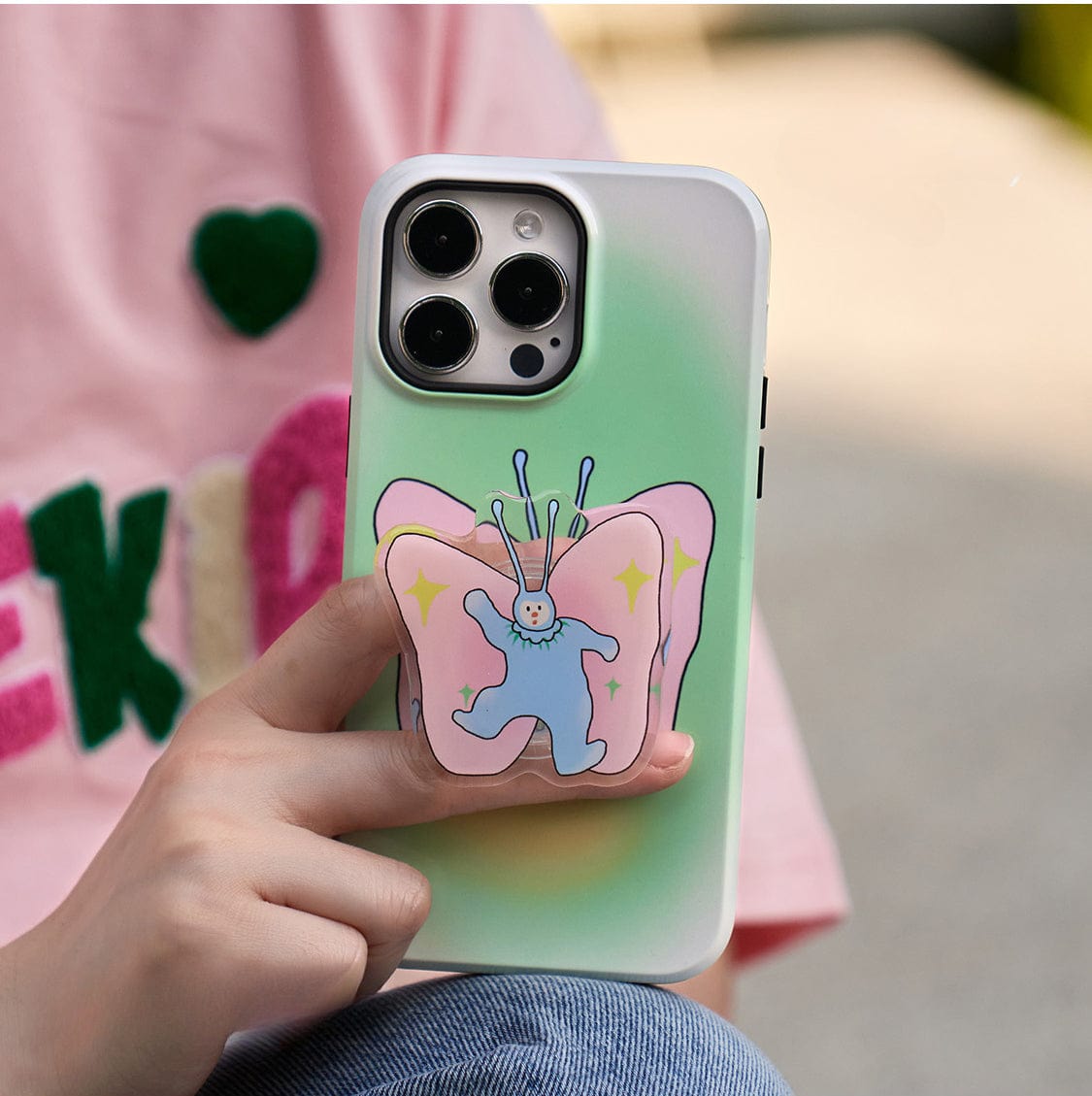 A person's hand holding the Golden Sunset MagSafe iPhone 16 Pro Max Case, featuring an elegant gradient design with a green and white color scheme. The case showcases a colorful sticker of a cartoon character resembling a butterfly with blue wings and a blue body. The individual is wearing a pink shirt adorned with vibrant, colorful lettering.