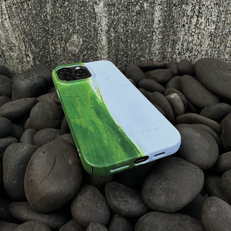 An iPhone 16 Pro Max with a Minimalist Green Brushstroke Case, featuring an artistic abstract design, is placed on smooth, dark river rocks. The phone's dual camera and charging port are visible, and the textured background suggests an outdoor or natural setting.