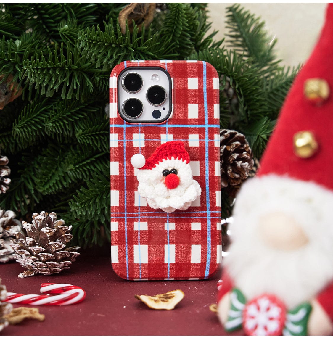 A smartphone with a three-camera setup is encased in a Festive Knitted MagSafe iPhone 16 Pro Max Case, featuring a small, fluffy Santa Claus face. It stands on a festive surface beside a pine tree branch with pine cones, a candy cane, and an out-of-focus gnome figurine wearing a red hat.