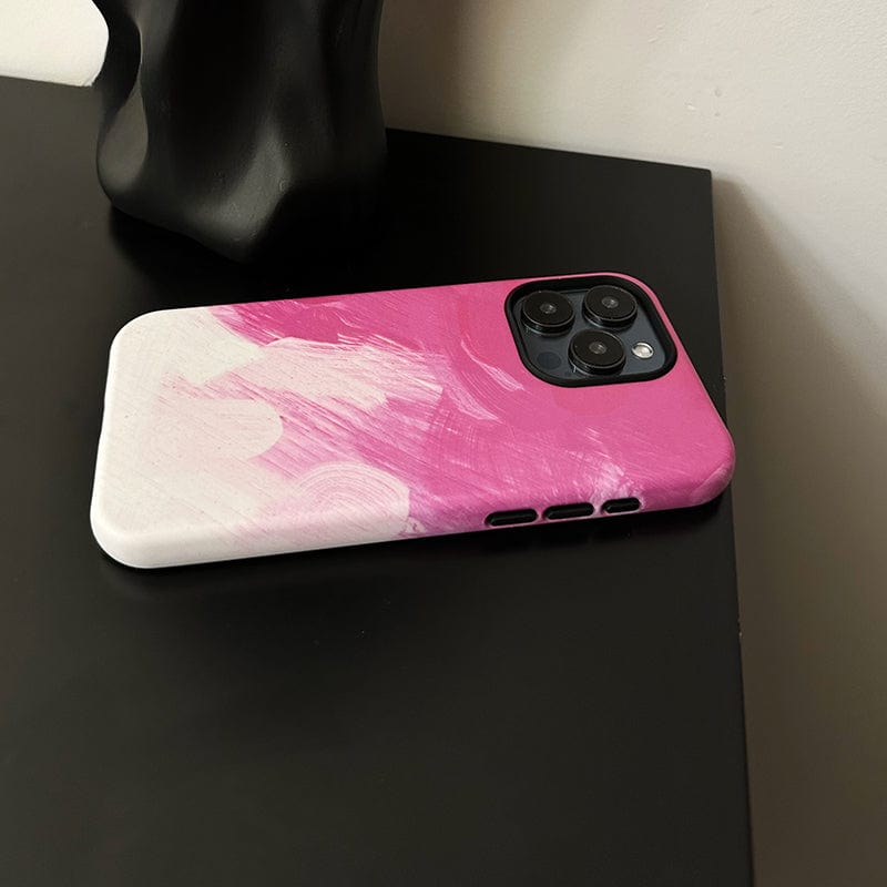 A smartphone encased in a Bold Pink Brushstroke iPhone 16 Pro Max Case - Artistic Design with Full Protection is lying on a black surface near a sculpted object. The case features two shades of pink blending into white abstract patterns. The phone has three camera lenses visible in the top corner.