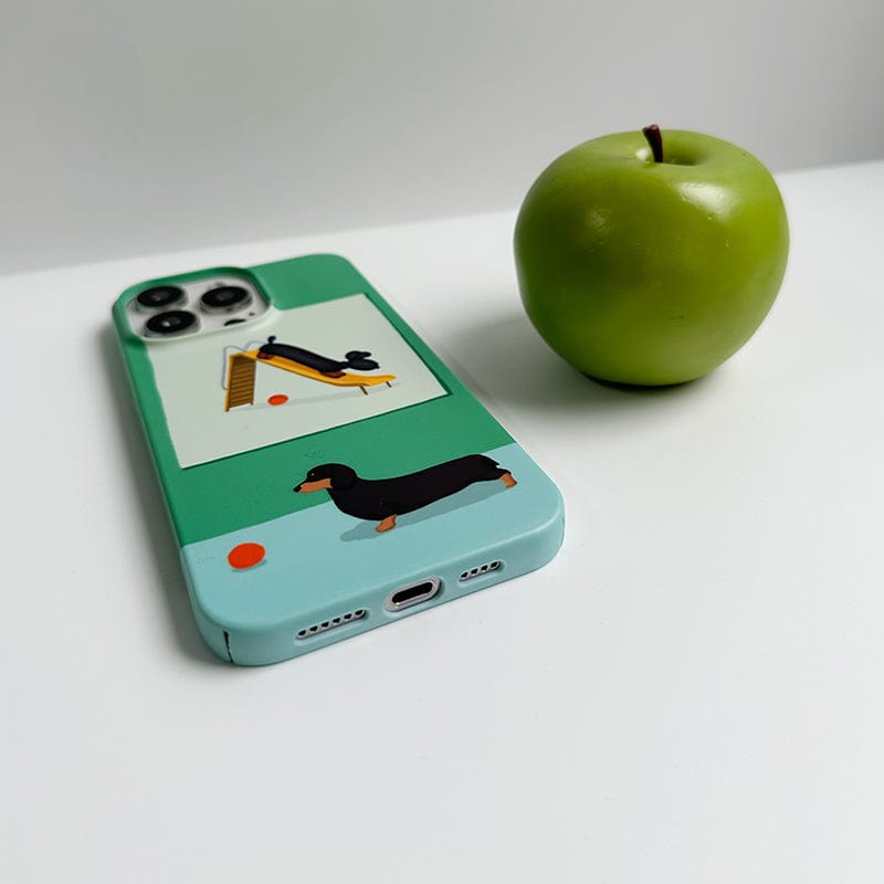 A smartphone with a Playful Dachshund iPhone 16 Pro Max Case featuring a cute green and blue design, resting on a white surface. Next to the smartphone is a green apple.