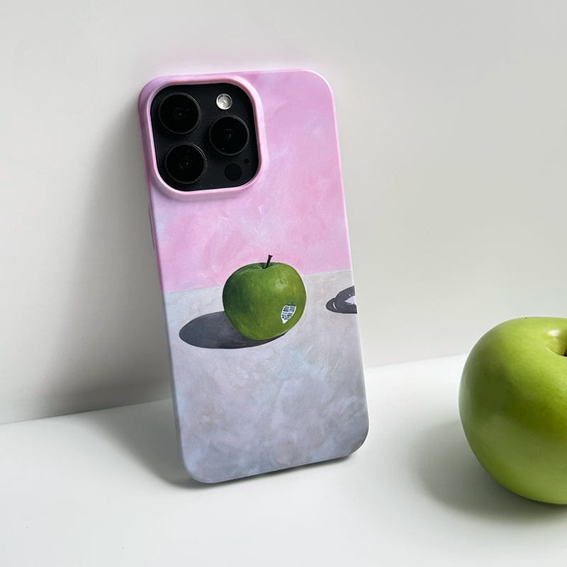 The Minimalist Green Apple iPhone 16 Pro Max Case features an artistic still life design, portraying a green apple casting a shadow on a soft pink background. To the right of the phone case, another green apple rests against a neutral backdrop.