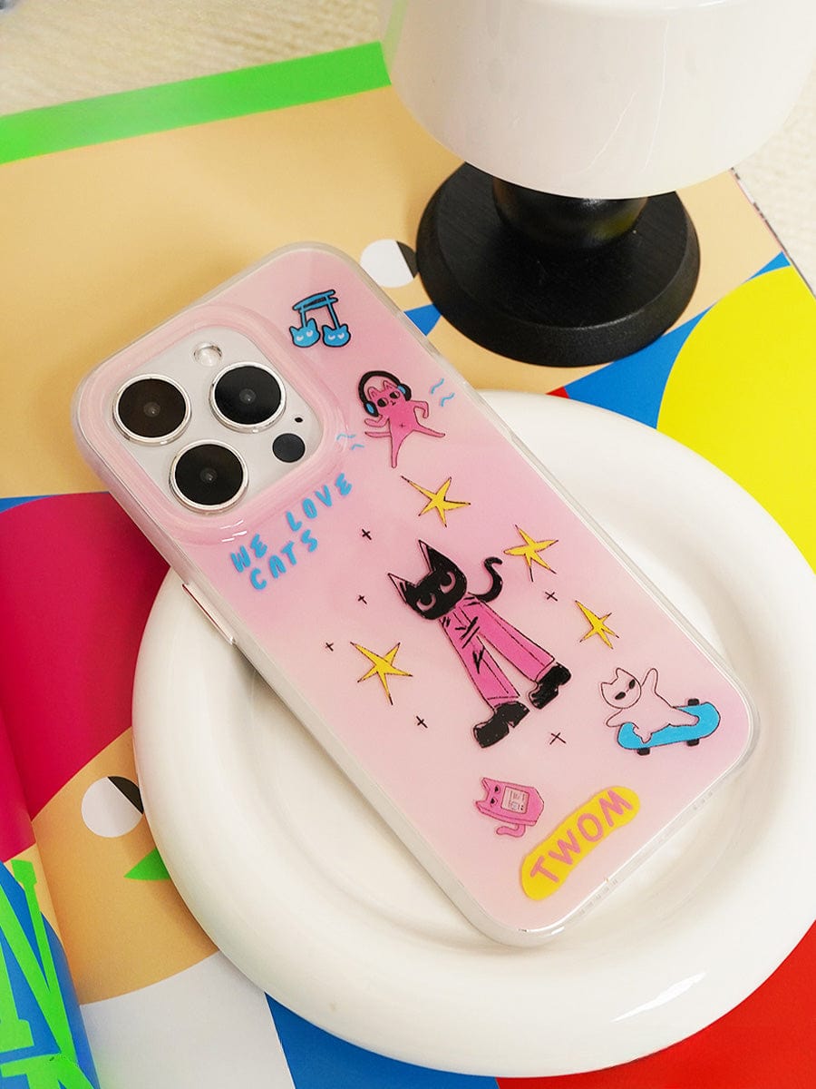 A We Love Cats iPhone 16 Pro Max Case, featuring a cute clear pink design adorned with illustrations of a black cat holding a book, a blue cat standing, a flying pink figure, stars, and the phrases "WE LOVE CATS" and "WOW!", rests on a circular white stand next to a black and white lamp.