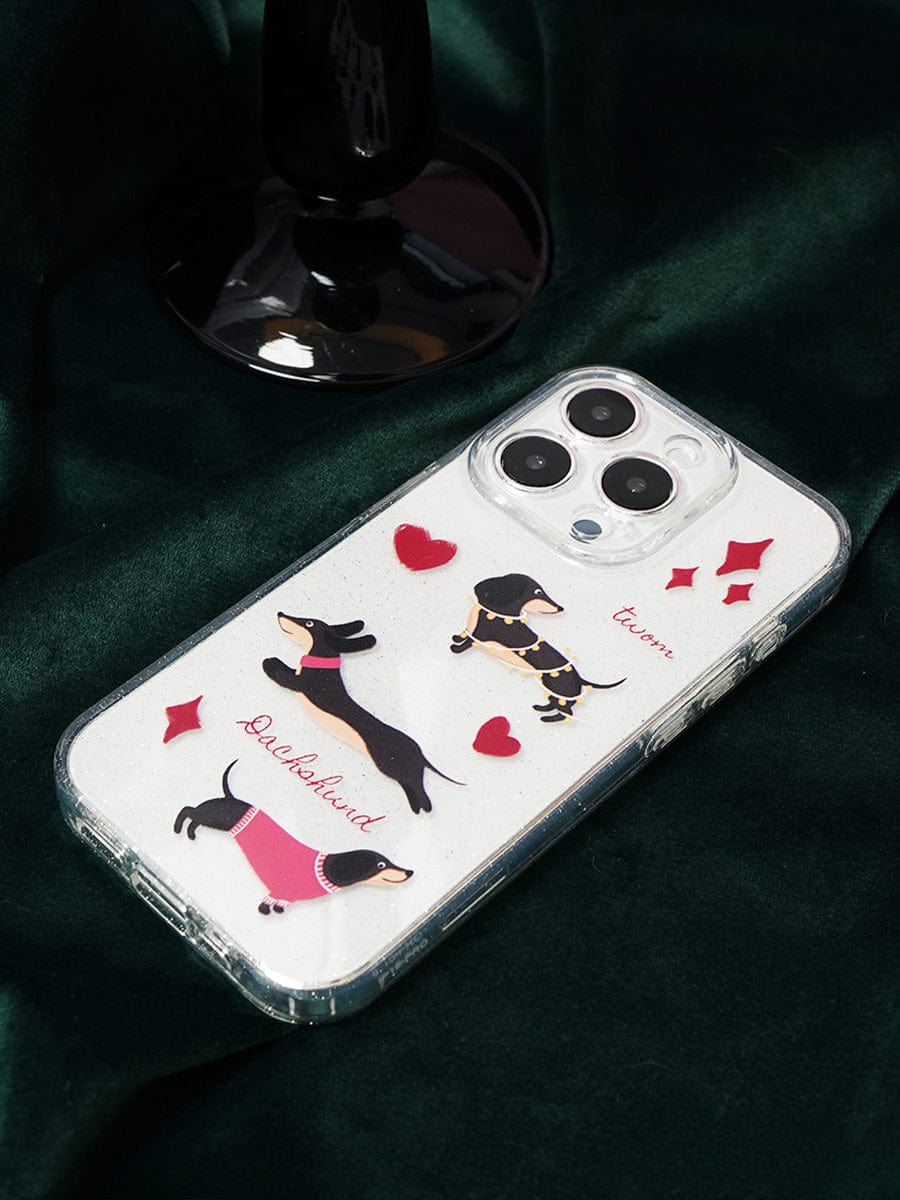 A smartphone adorned with the "Dachshund Love iPhone 16 Pro Max Case | Clear Sparkle Design | Cute All-Inclusive Protective Cover" is resting on dark green fabric. The clear case showcases images of three dachshunds dressed in clothes, surrounded by red hearts and diamonds. The word "Dachshund" is elegantly displayed alongside one of the dachshunds.