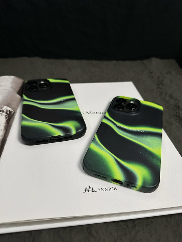 Two smartphones with matching Abstract Wave iPhone 16 Pro Max cases, each showcasing a sleek black and green gradient design with the "Happier Than Ever" quote, are placed on an open book. The cases feature a flowing green pattern that contrasts against the black background. The partially visible book beneath them appears to be titled "ANNICE".