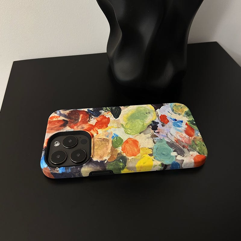 An Abstract Paint Palette iPhone 16 Pro Max Case - Artistic and Protective lies on a black surface. The design resembles a painter's palette with various splotches of vibrant colors like red, green, blue, and yellow. The background shows part of a black sculptural object.