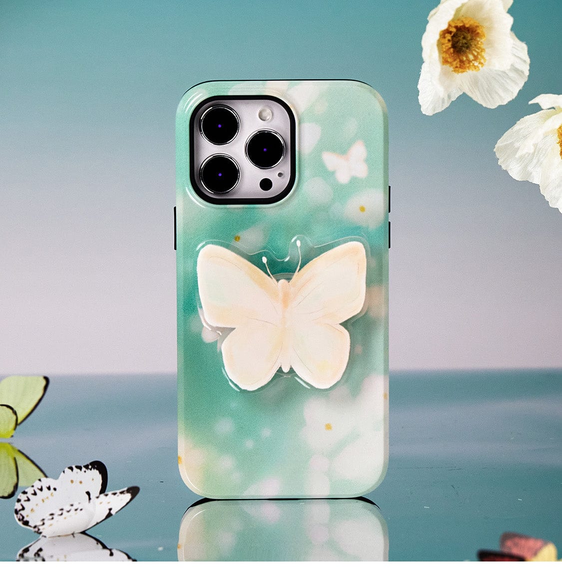 The Butterfly Blossom MagSafe iPhone 16 Pro Max Case, an all-inclusive protective cover with an elegant floral design and built-in stand, showcases a large cream-colored butterfly at the center on a soft green background. This smartphone accessory is displayed on a reflective surface with white flowers nearby.