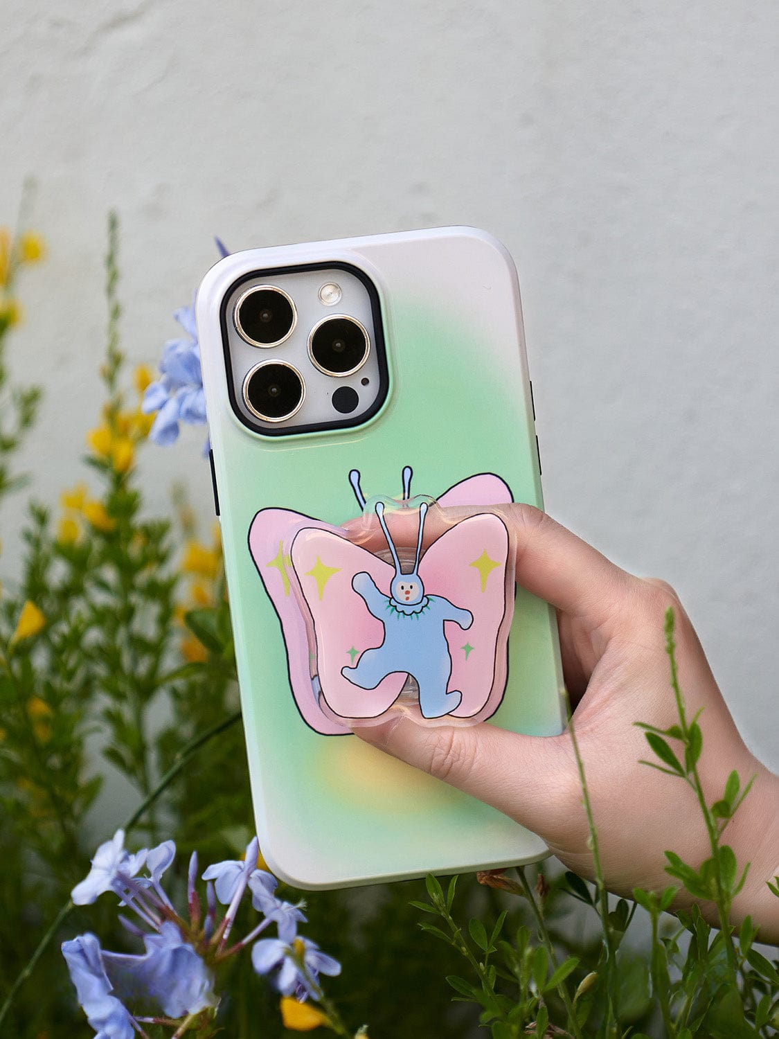 A hand holds the Golden Sunset MagSafe iPhone 16 Pro Max Case, featuring an elegant gradient design from green to pink. The whimsical butterfly design on the case has a blue center with a happy face and is surrounded by yellow stars. Green plants and blue flowers decorate the background, completing this all-inclusive protective cover with a built-in ring stand.