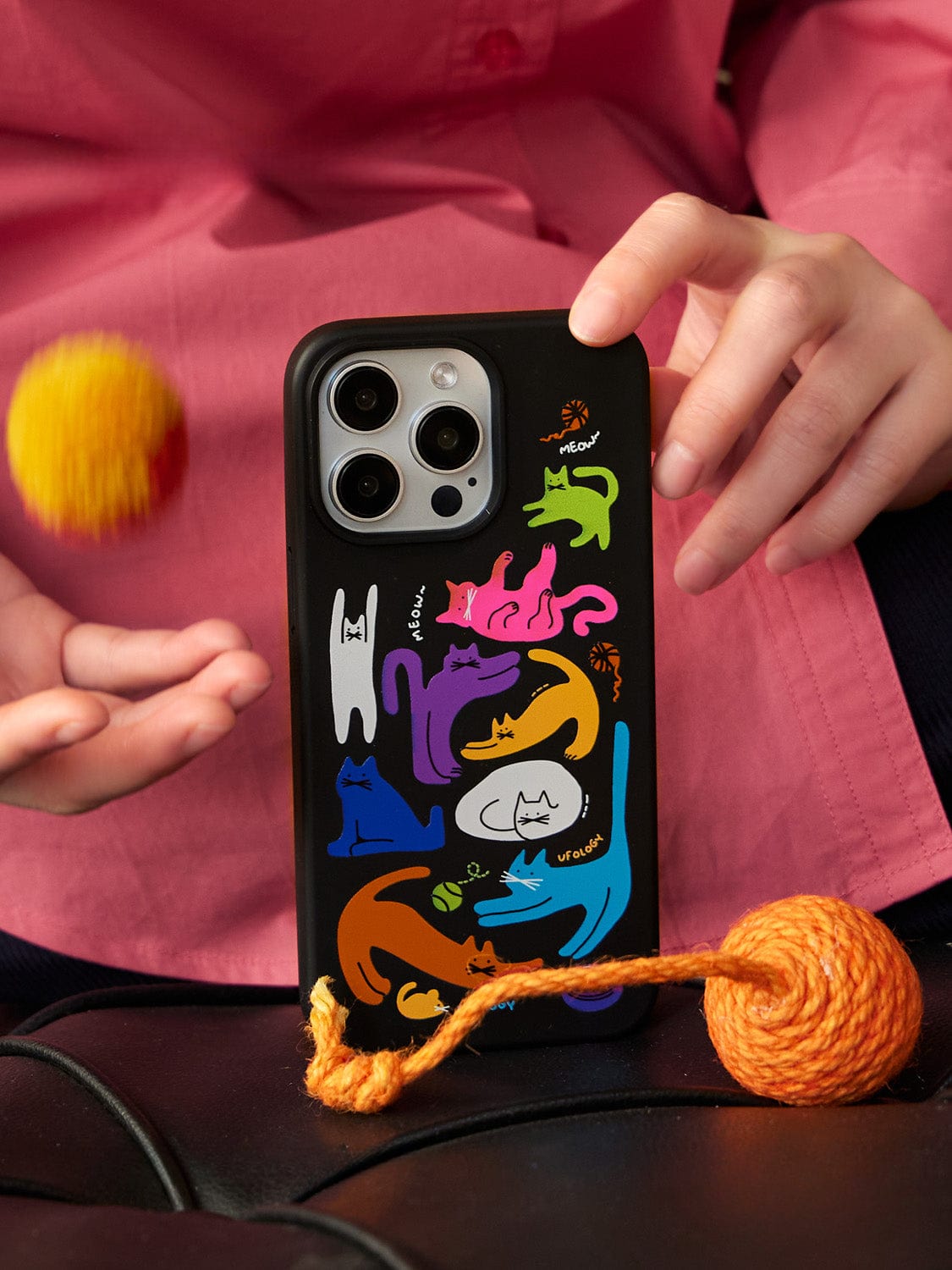 A person in a pink shirt is holding an iPhone 16 Pro Max with a Colorful Cats MagSafe case, featuring a vibrant cat design and built-in stand, while also tossing an orange ball of yarn.