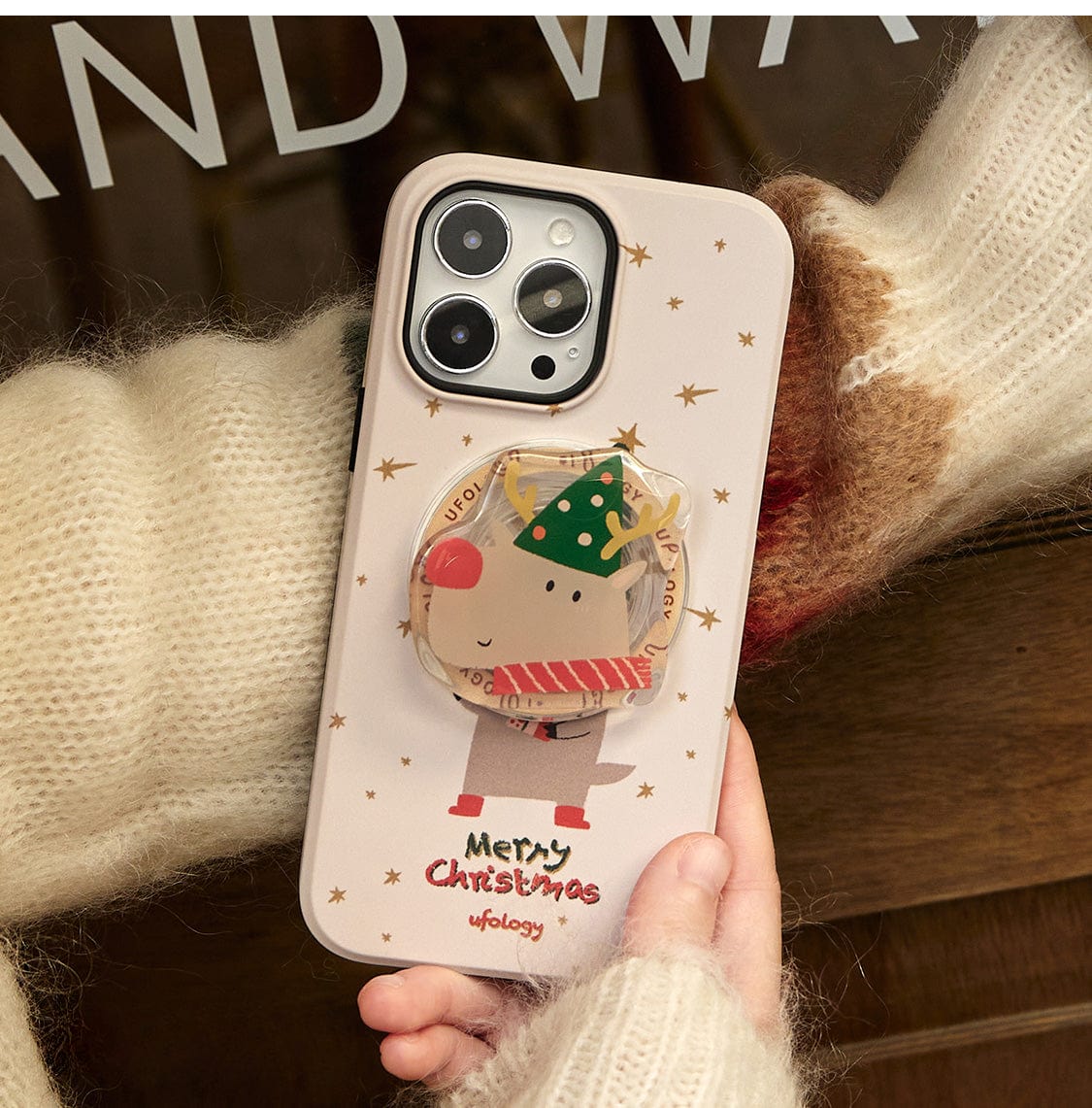 A hand wearing a fuzzy, cream and brown sweater holds a phone with a Rudolph Joy MagSafe iPhone 16 Pro Max Case, featuring a festive reindeer design and built-in stand, against a reflective surface.