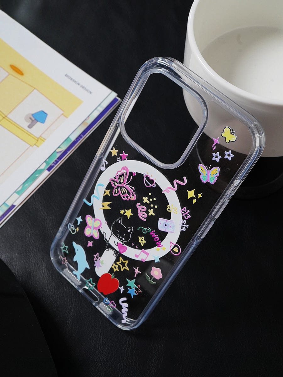 A Whimsical Dreams MagSafe iPhone 16 Pro Max Case, featuring a transparent fun design with colorful cartoon animals, butterflies, and hearts, lies on a black surface next to a white cup and an open book.