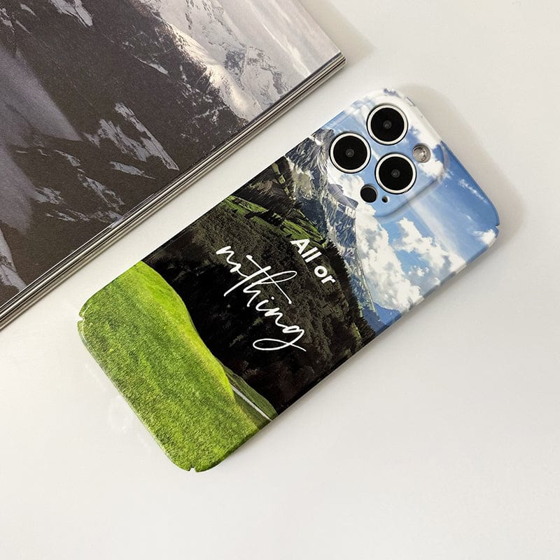 Close-up of the Mountain Landscape iPhone 16 Pro Max Case, displaying a scenic landscape with green hills, a partly cloudy sky, and the motivational quote "All or Nothing" in cursive. The phone rests on a white surface beside a book featuring a black and white mountain photograph.