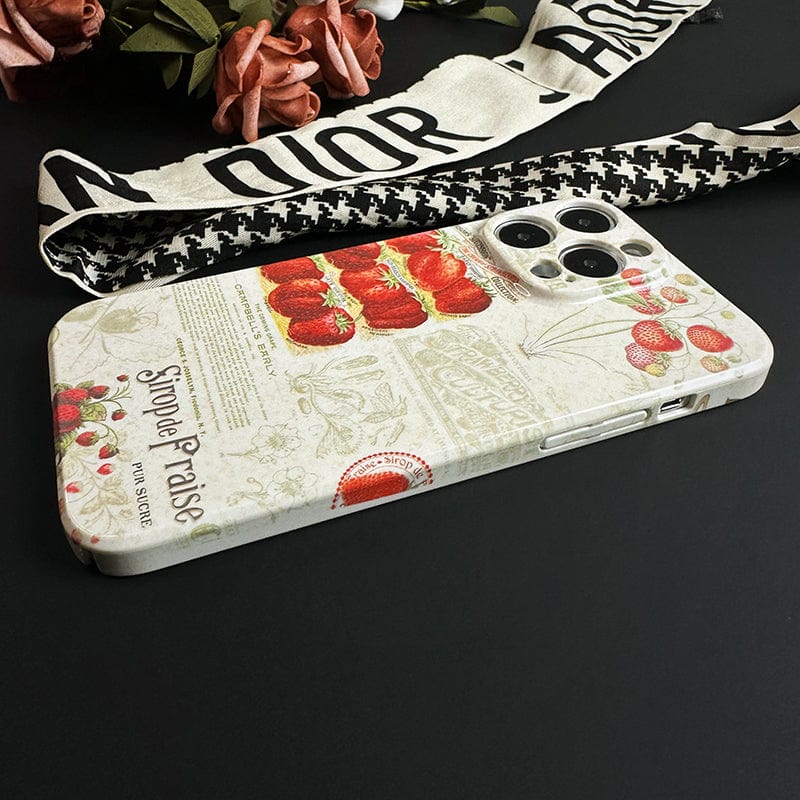 A smartphone with a Vintage Strawberry Print iPhone 16 Pro Max Case - Elegant and Protective rests on a black surface. Nearby are faux red roses and a white Dior cloth with black text. The phone case's design, featuring illustrations and French script, lends a nostalgic charm.