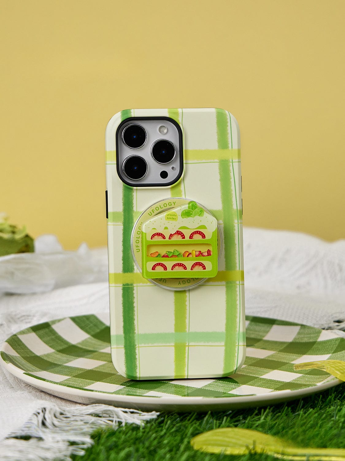 An iPhone adorned with the Fresh Picnic MagSafe iPhone 16 Pro Max case, featuring a green plaid design and integrated sandwich stand, alongside a sushi-themed circular phone grip is showcased on a matching green plaid plate against a yellow backdrop.