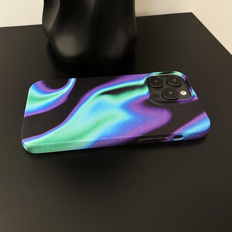 The Iridescent Waves iPhone 16 Pro Max Case, featuring a sleek and futuristic design with a black, green, and purple swirling pattern, rests on a black surface. The phone's camera lenses are visible in the top left corner of the case. In the background is an abstract-shaped black object.