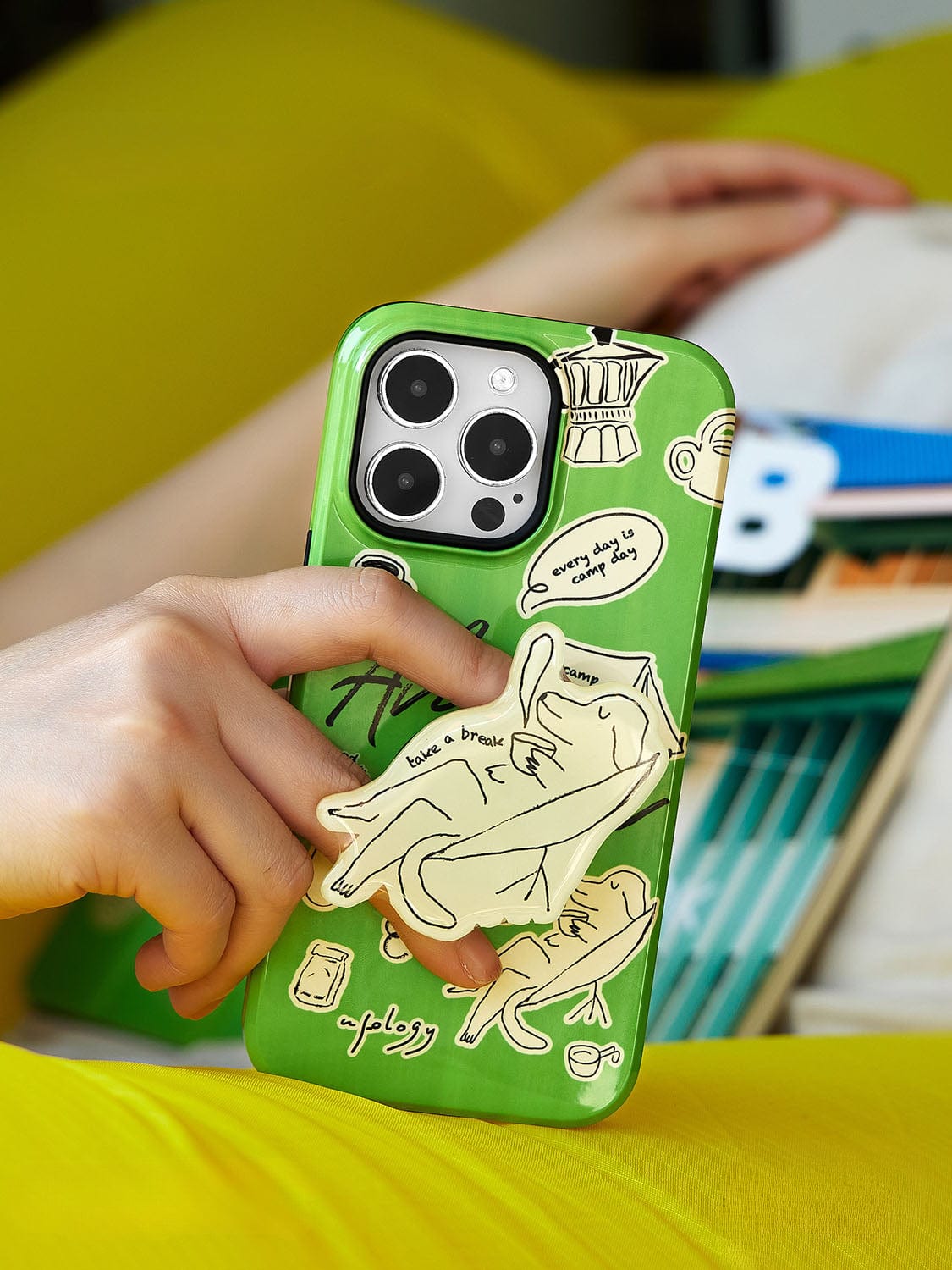 A hand holds a green Lazy Day Elephant MagSafe iPhone 16 Pro Max Case, adorned with various stickers, including a cartoon hand and text. The background features a colorful book and a yellow cushion, perfectly complementing the playful design of the all-inclusive protective cover with a built-in stand.