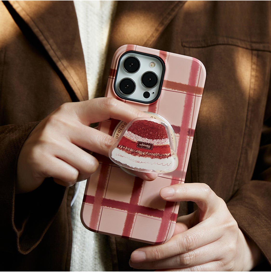 A person wearing a brown coat holds a smartphone with a Cozy Plaid MagSafe iPhone 16 Pro Max case that features a warm winter hat design and includes an all-inclusive protective cover, along with a pop socket showcasing a strawberry cake design.