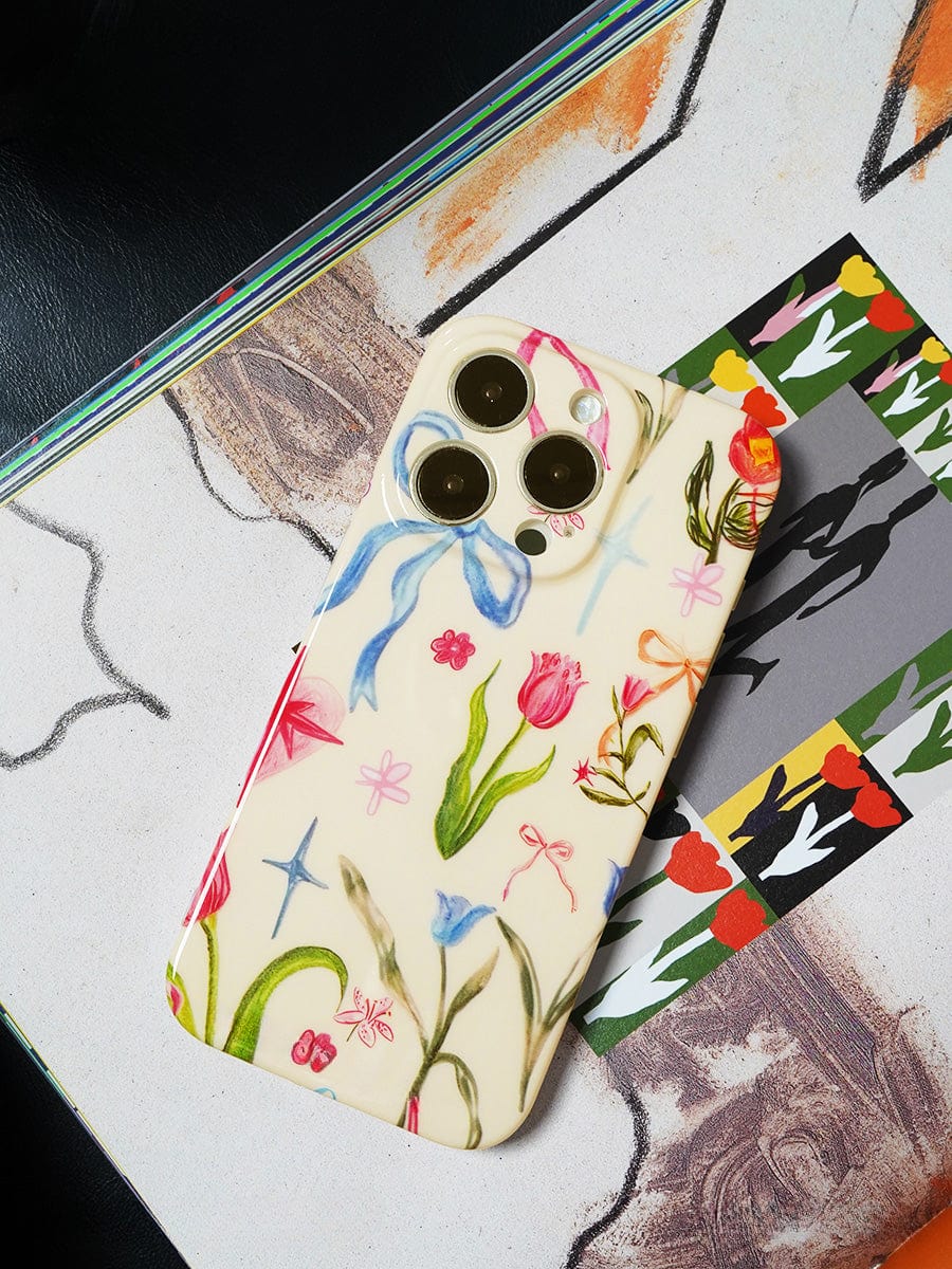 A smartphone with a Floral Dreams iPhone 16 Pro Max Case showcasing a colorful botanical design is placed on a vibrant piece of paper. Three camera lenses are visible on the back of the phone.