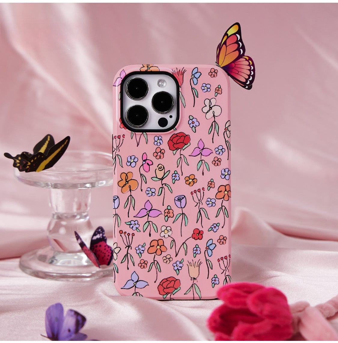 The Blossom Butterfly MagSafe iPhone 16 Pro Max Case, featuring a delightful floral pink design, rests on a pink fabric surface. Several decorative butterflies adorn and surround the phone case, enhancing its whimsical appeal. In the background, an empty clear glass butterfly stand completes the scene.