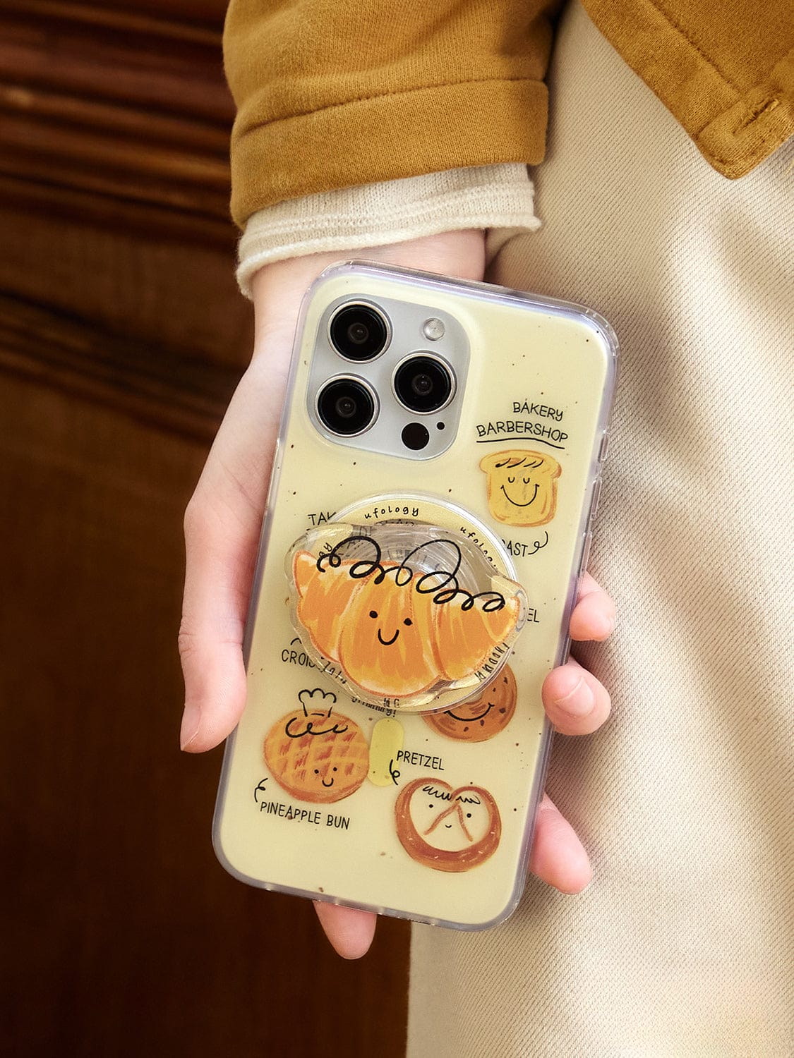 A person is holding an iPhone with the Bakery Friends MagSafe iPhone 16 Pro Max Case, which has a transparent design featuring a smiling bread cartoon and various pastries like pretzel, pineapple bun, croissant, muffin, and more. The all-inclusive protective cover also includes text referencing a bakery and breadshop along with a central Bun Stand pop socket.