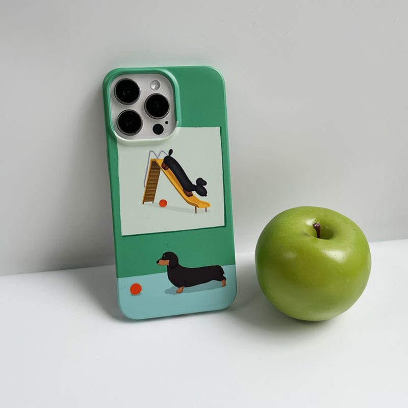 A Playful Dachshund iPhone 16 Pro Max Case featuring a cute green and blue design with illustrations of a dachshund playing on a slide and next to an orange ball is propped against a white background. Next to the phone case is a green apple.