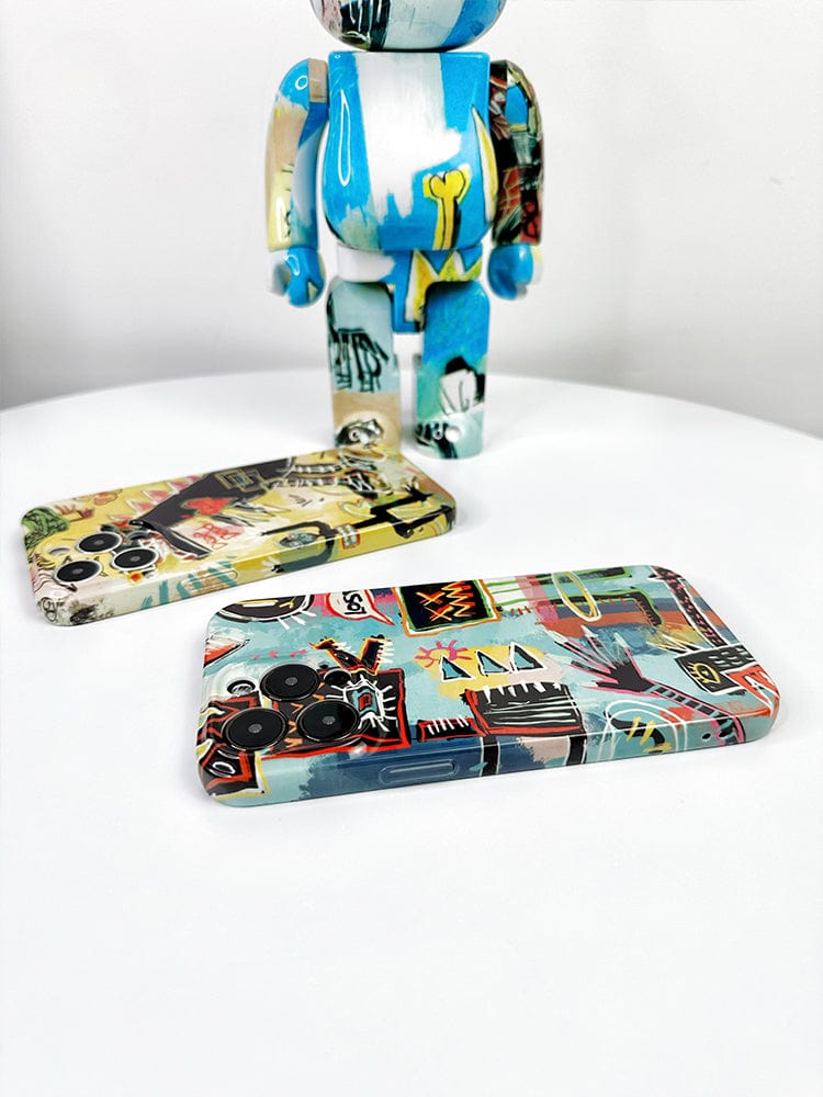 An artistic action figure with a blue and white abstract design stands on a white surface. In front of it lie two cases from the Urban Graffiti iPhone 16 Pro Max Case Set - Bold Street Art Collection, each adorned with colorful, abstract patterns featuring various shapes and symbols.