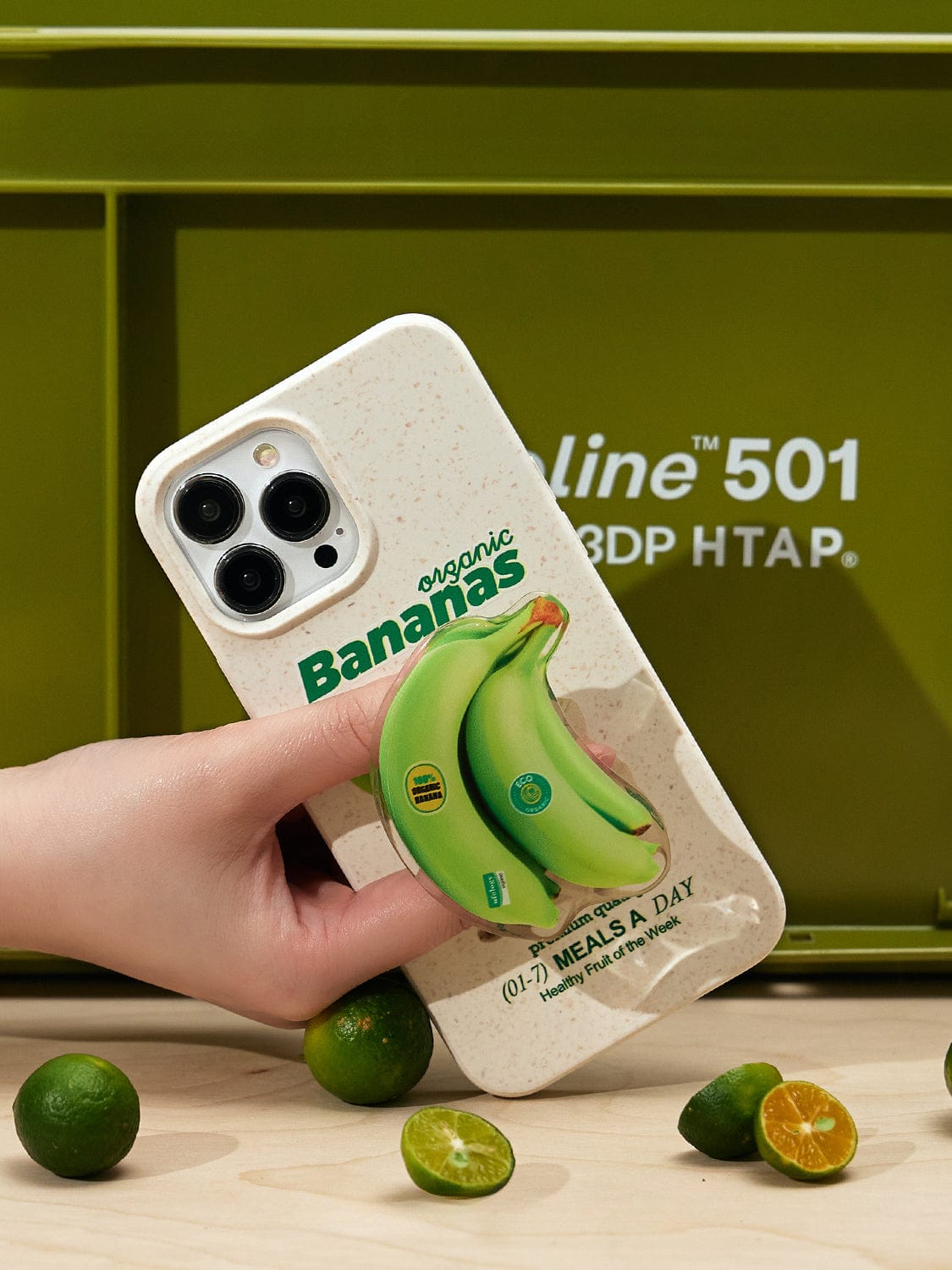 A hand is holding the Organic Bananas MagSafe iPhone 16 Pro Max Case, which showcases a fun fruit design featuring a 3D image of a green banana and the text "Organic Bananas." In the background, there's a green box and several small limes, with one lime cut in half.