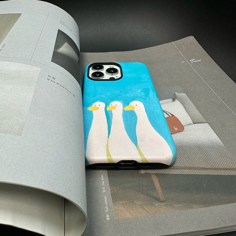 A smartphone with a Whimsical Three Ducks iPhone 16 Pro Max Case - Playful Blue & Yellow Design, Ultra-Slim, Full Protection featuring artwork of three white ducks standing in a row lies on an open magazine. The magazine displays images and text, and the background is a dark surface.