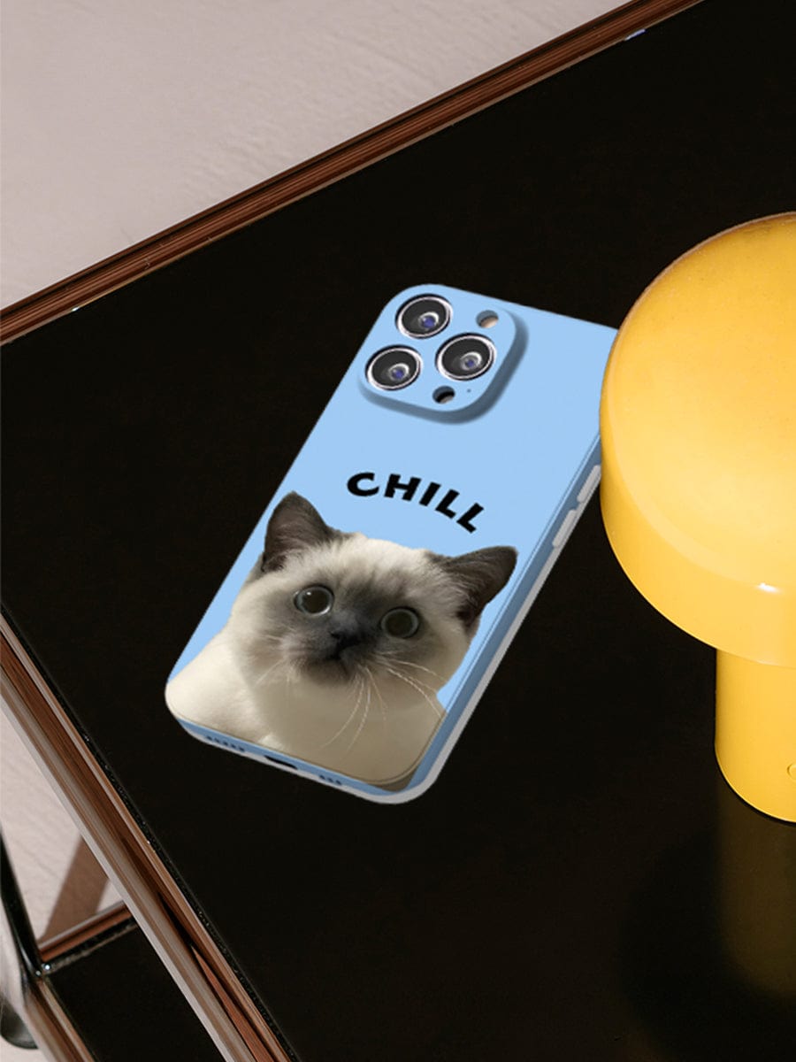 An iPhone 16 Pro Max, encased in the Chill Cat iPhone 16 Pro Max Case featuring an adorable kitten design and the word "CHILL," is placed on a black surface next to a yellow object. The phone's three camera lenses are clearly visible at the top.