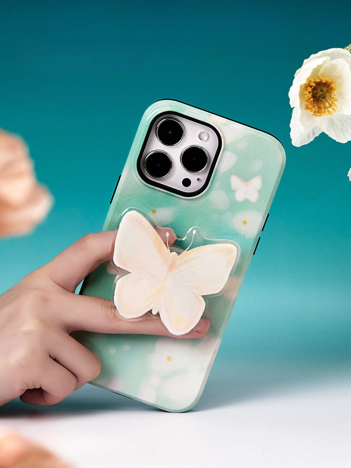 Hand holding an iPhone 16 Pro Max with a Butterfly Blossom MagSafe case, featuring a green background and white butterflies, against a teal backdrop adorned with flowers.