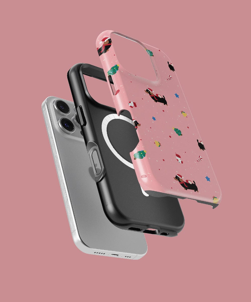 Three iPhone cases hover in the air, providing robust protection. They come in different shades: gray, solid black, and a delightful pink adorned with the Christmas Dachshund pattern that exudes personality. The MagSafe-compatible design ensures clear visibility of the rear camera. All this is set against a soft pink backdrop.